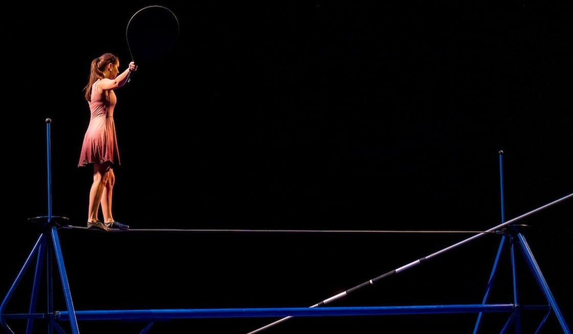 An aerial dance student crosses a tight rope on a dramatically lit stage.