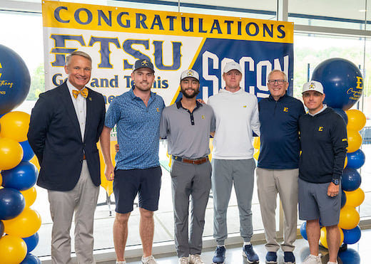 The men's golf team poses with ETSU President Brian Noland and Athletic Director Doc Sander in front of a SoCon Championship banner. 