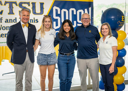 The women's golf team poses with ETSU President Brian Noland and Athletic Director Doc Sander in front of a SoCon Championship banner. 