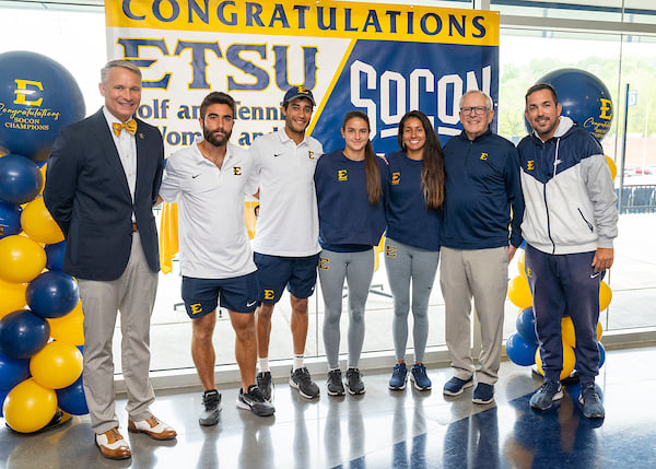 The men's and women's tennis teams pose with ETSU President Brian Noland and Athletic Director Doc Sander in front of a SoCon Championship banner. 