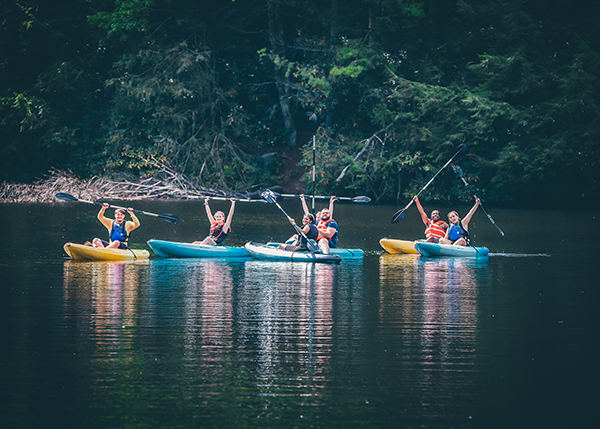 ETSU students sitting in blue and gold kayaks raise their paddles in the air in celebration. The students are on a lake with lush green foliage in the background. 