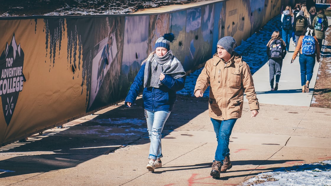 students bundled in winter clothing walk along the campus center construction site. Lingering piles of snow are visible. 