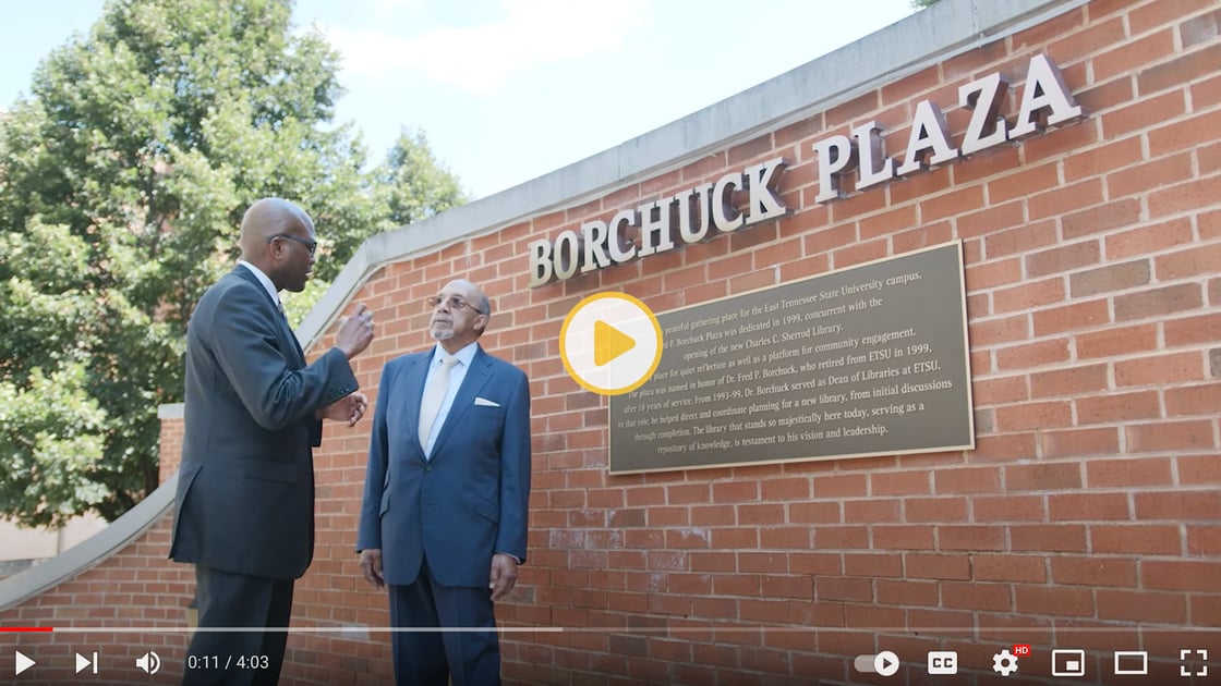 A screenshot of a video. Two men are shown talking in front of an arched brick wall, which bears the name “Borchuck Plaza” in elegant bronze letters.  