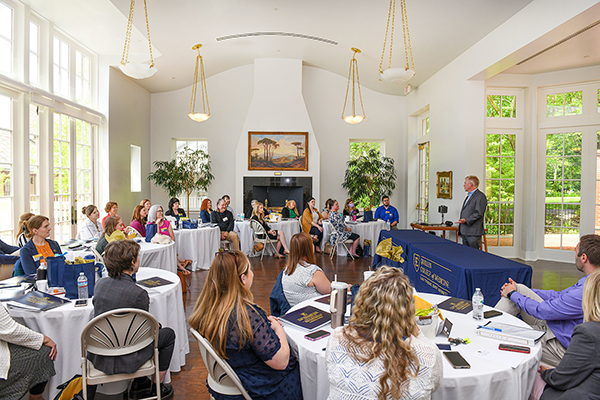 Students and faculty sit at tables in a meeting room listening to a presentation.