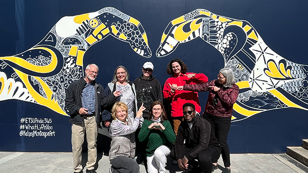 Team members from the Office of University Marketing and Communications pose with illustrator Kelsey Montague in front of a newly created mural on campus. The mural depicts two hands forming the shape of a heart. Within the hands are sketched symbols and patterns, some representing East Tennessee and ETSU. Examples include the ETSU Carillon, rhododendrons, mountains, the Ballad Health Athletic Center (Mini-Dome), and the numerals 1911, indicating the year ETSU was founded.