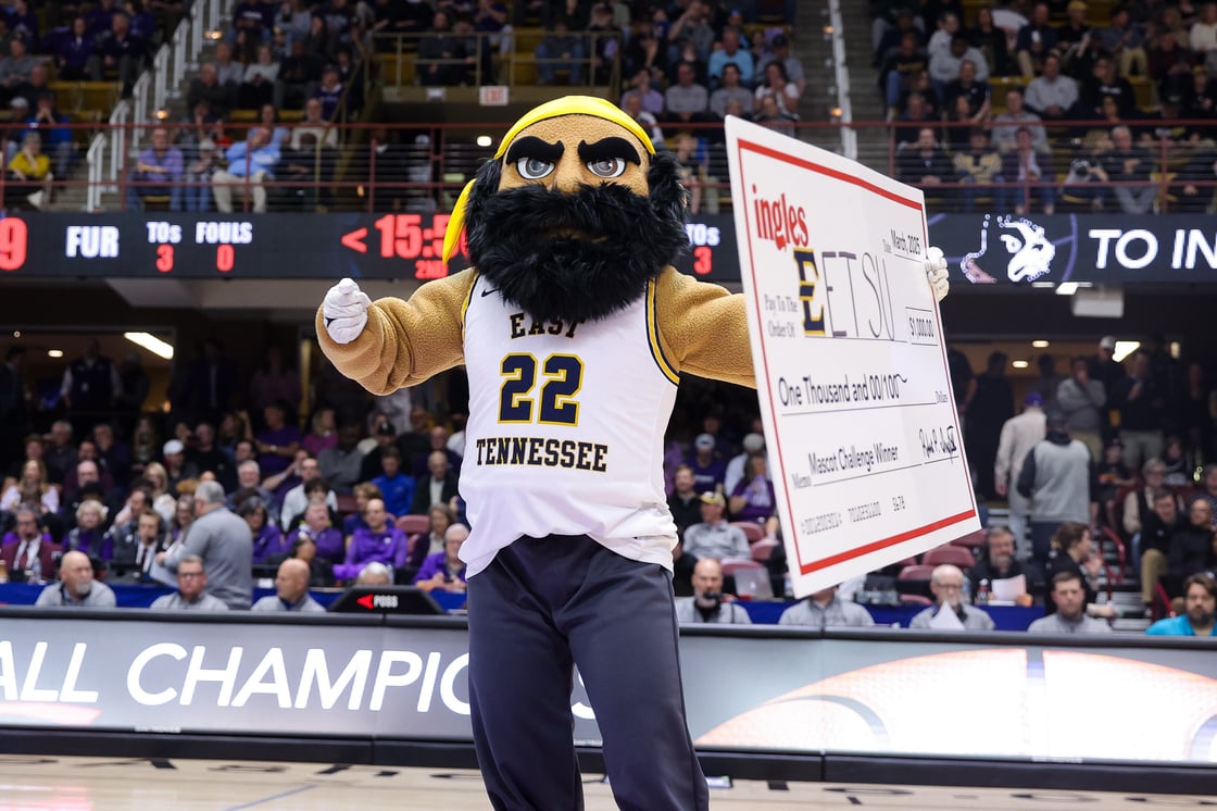 ETSU's mascot, Bucky, stands on a basketball court holding a giant check for $1,000, awarded for winning the Ingles Mascot Challenge. Bucky wears a white and gold East Tennessee jersey with the number 22, striking a victorious pose in front of a crowd of spectators in the arena.