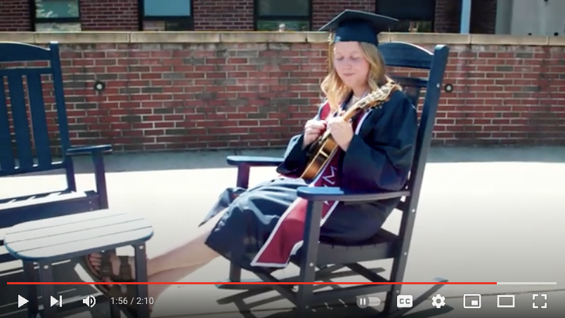Lydia Hamby sits in a rocking chair on a sunny day on campus. She is wearing her cap and gown and playing her mandolin.  