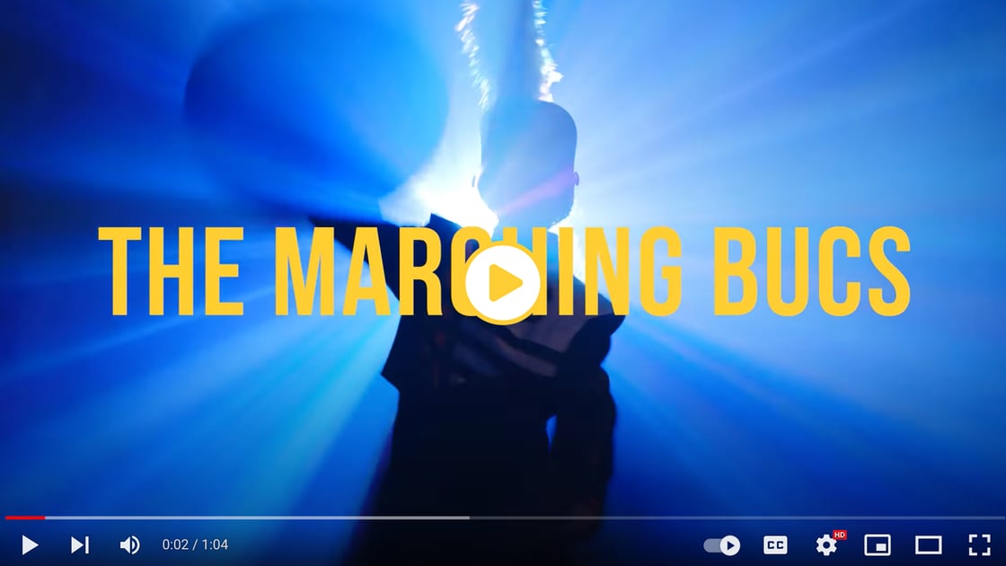 A screenshot of a video featuring a marching band member silhouetted against blue lights. Text over the frame reads “Marching Bucs.”