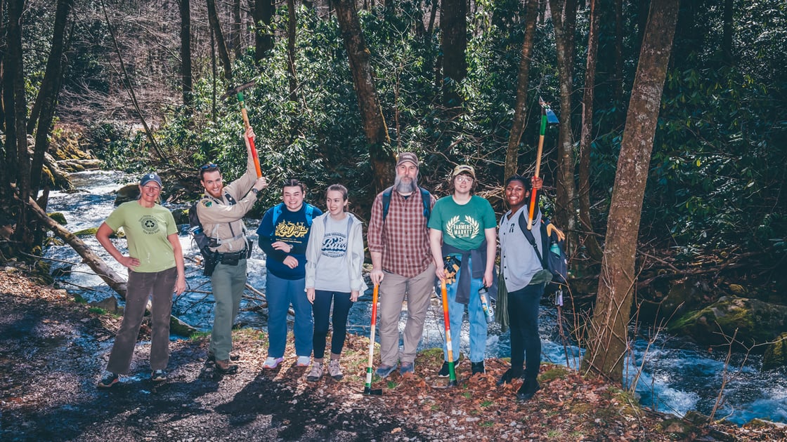Several smiling students pose with shovels in front of a beautiful stream in the forest. 