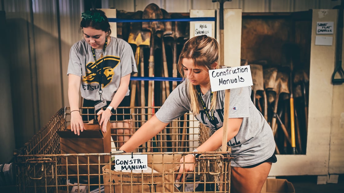Two students wearing ETSU gear sort materials in a warehouse. 