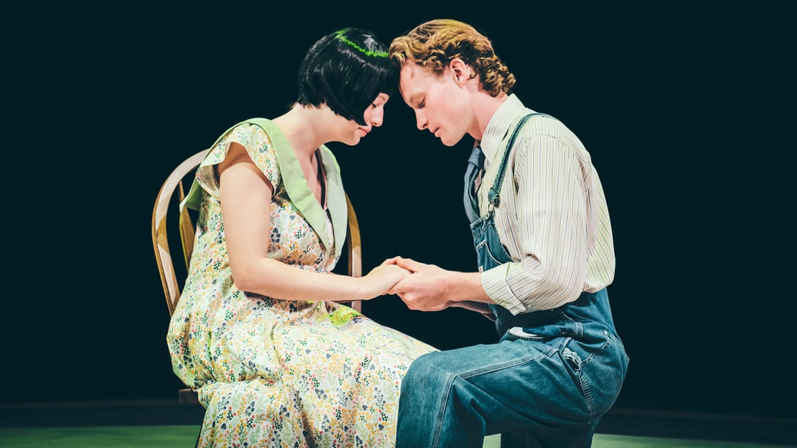 A man in overalls and a woman in a flowered dress lean against each other, forehead to forehead. Their eyes are closed and they are holding hands. 