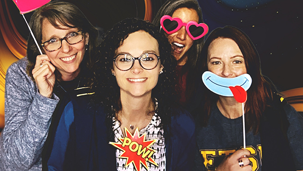 Four university staff members pose holding props, including a fake mustache and a princess crown.