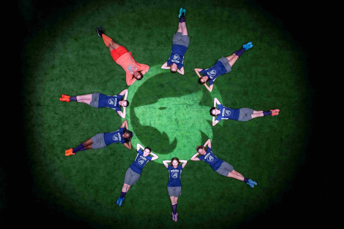 An overhead shot of a soccer team in blue and gray uniforms lying on artificial turf in a circular formation around a wolf logo, with the goalkeeper dressed in orange positioned at the top.